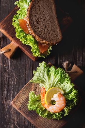Sandwich with whole wheat bread, fresh salad, shrimp and salted salmon on little wooden cutting board over old wooden table. Rustic style. Top viewの写真素材