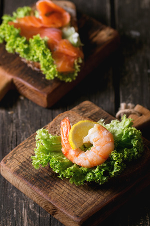 Sandwiches with whole wheat bread, fresh salad, shrimp and salted salmon on little wooden cutting board over old wooden table. Rustic style.の写真素材