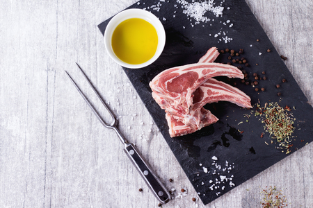 Raw lamb chops with salt, pepper, dry herbs and bowl of olive oil on black slate board over white wooden table. Meat fork near. Top view. With copy space at leftの写真素材