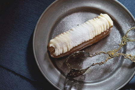Single Eclair with white butter cream in vintage metal plate with cake tongs. Dark rustic style. Day light. Top viewの写真素材