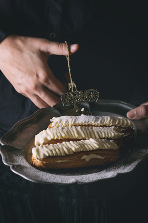 Woman's hands take by cake tongs from vintage metal plate Eclairs with white butter cream. Dark rustic style. Day light. In retro filter effectの写真素材