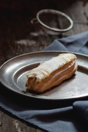 Single Eclair with white butter cream in vintage metal plate. Dark rustic style. Day light. Outside...