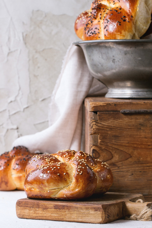 Heap of sweet round sabbath challah bread with white and black sesame seeds in vintage metal bowl on wooden chest and on small cutting board over white table with plastered wall at background.の写真素材