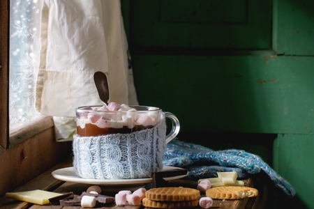 Glass cup of Hot chocolate with marshmallows in knitted cup holder with cookies, chopping chocolate and mittens over wooden window sill near frozen window in sunny day. Rustic style.の写真素材