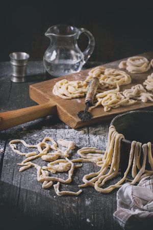 Fresh homemade pici pasta on wood chopping board over old wooden table with flour, copper bowl, rolling-pin and galss jug of water. Dark rustic style with retro filter effect. See process seriesの写真素材