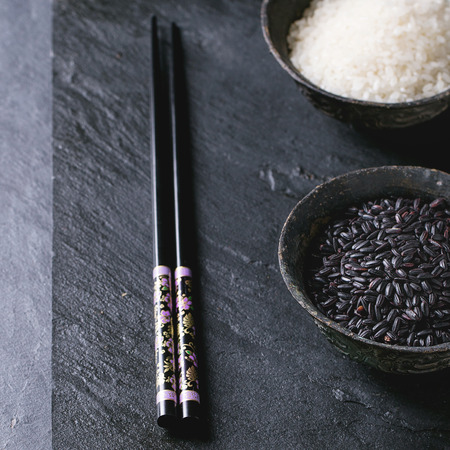 Black and white rice in old metal china bowls with black chopsticks over black slate background. Square image with selective focusの写真素材