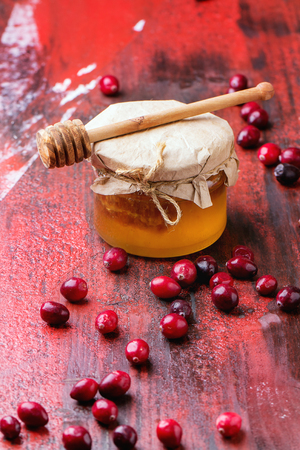 Glass jar of liquid honey with honeycomb inside, and fresh cranberries over red and black wooden table.の写真素材