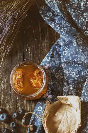 Open glass jar of liquid honey with honeycomb inside, fresh blueberries and bunch of dry lavender over old wooden table with blue textile rag. Dark rustic style. Top view. With retro filter effectの写真素材