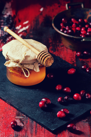 Glass jar of liquid honey with honeycomb inside, and fresh cranberries on black slate board over red and black wooden table. With retro filter effectの写真素材