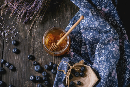 Open glass jar of liquid honey with honeycomb and honey dipper inside, fresh blueberries and bunch...