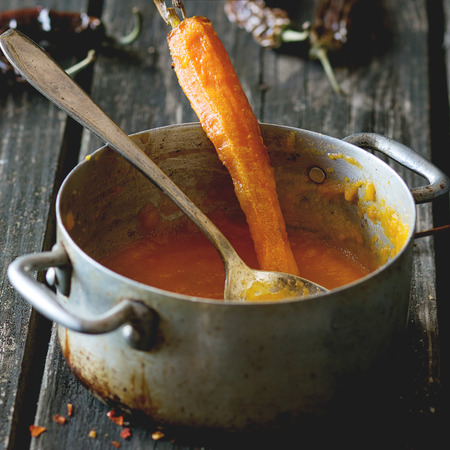 Aluminum pan with remains of carrot cream soup with whole baked carrot and red hot chili pepper. Over old wooden table with vintage spoon and dry peppers. Dark rustic style. Square image with selective focusの写真素材