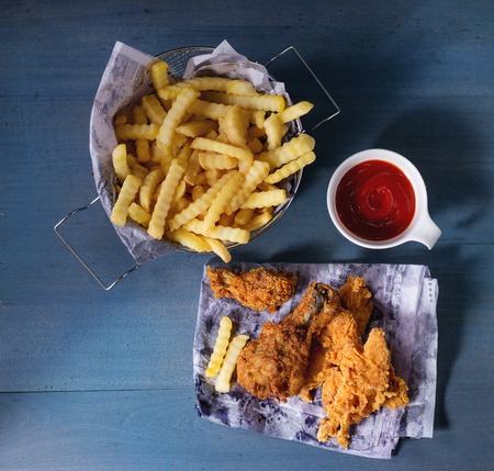 Chicken fries strips and legs on paper with basket of  French fries and bowl of ketchup sauce over blue wooden table. Top view. Square imageの写真素材
