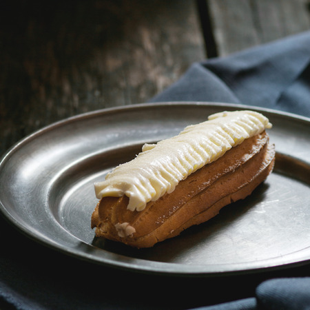 Single Eclair with white butter cream in vintage metal plate. Dark rustic style. Day light. Outside...