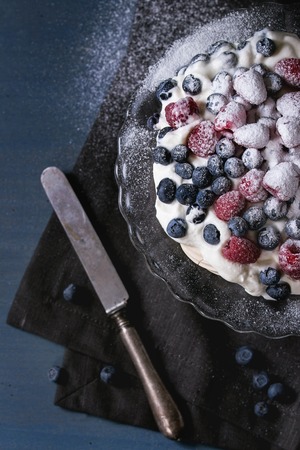 Vintage cake stand with Meringue dessert Pavlova with sugar powder, fresh blackberries and raspberries. Over blue wooden table with old knife and textile napkin. Dark rustik style. Flat layの写真素材