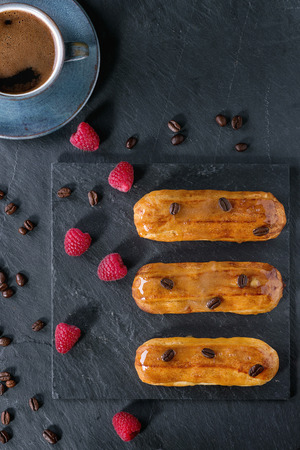 Coffe eclairs with cup of fresh black coffee, raspberries and coffee beans on black slate board over blue wooden surface. With space for text. Flat layの写真素材