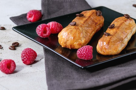 Black squate plate with Coffe eclairs with fresh raspberries and coffee beans on gray textile napkin...