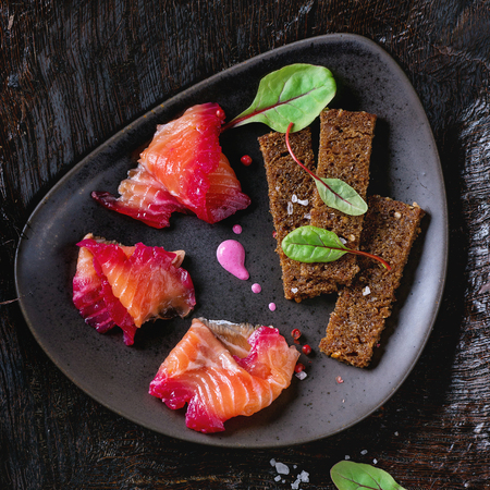 Triangular ceramic plate with Sliced salmon filet, salted with beetroot juice, served with whole wheat toasts, salad leaves and beetroot sauce over black surface. Flat lay. Square imageの写真素材