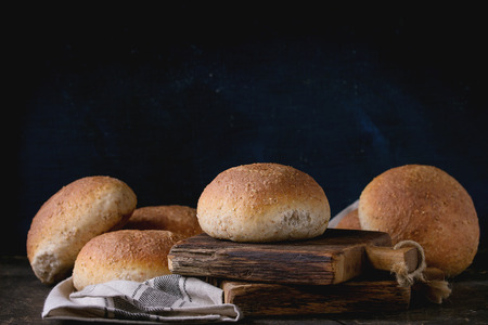 Fresh baked wholegrain buns on gray kitchen towel over small wooden chopping board with black background.の写真素材