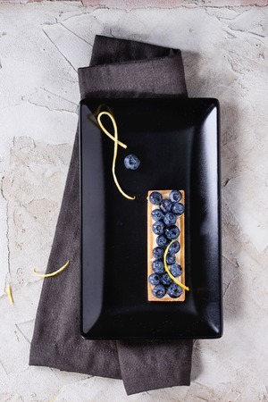 Square Lemon tartles with fresh blueberries, served on black square plate with lemon zest and textile napkin over gray plastered surface. Flat lay. With space for textの写真素材