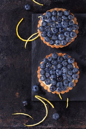 Two Lemon tartlets with fresh blueberries, served on black stone slate with lemon and lemon zest over black background. Top view. With space for textの写真素材