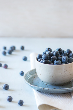 Spotted blue ceramic cup of blueberries with saucer at white textile napkin over wooden table. Rustic style. Natural day lightの写真素材