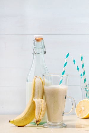 Glass of fresh Banana smoothie with retro cocktail tube, served with bottle of milk, banana and half of lemon over white wooden table. Rustic styleの写真素材