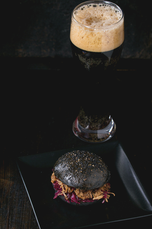 Black homemade burger with beef stews and red cabbage served on black square plate with glass of dark beer over wooden table with black background.の写真素材