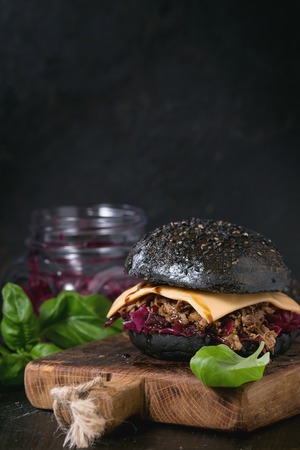 Black burger with beef stews, cheese, red cabbage and balsamic sauce served on small wooden chopping board with fresh basil over wooden table with black backgroundの写真素材