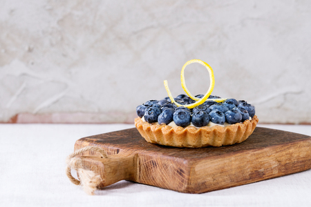 Lemon tartlet with fresh blueberrie and lemon zest, served on small wooden chopping board over white table.の写真素材
