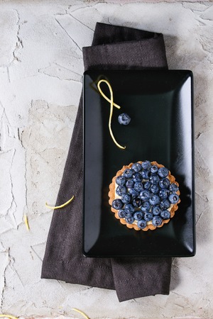 Lemon tartles with fresh blueberries, served on black square plate with lemon zest and textile napkin over gray plastered surface. Flat lay. With space for textの写真素材
