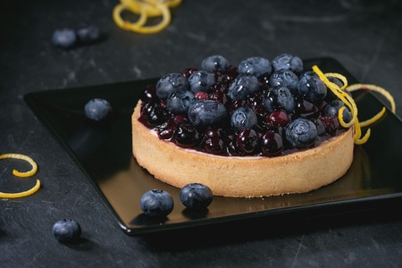 Round Lemon Tart with fresh and cooked blueberries, served on black square plate with lemon zest over black background.の写真素材