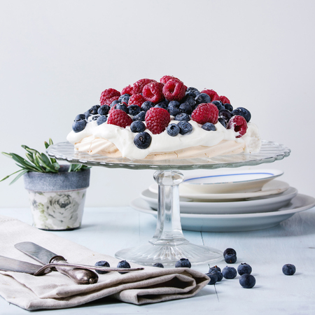 Vintage cake stand with Meringue dessert Pavlova with fresh blackberries and raspberries. Over blue wooden table with old tableware, textile napkin and snowdrops flowers.の写真素材
