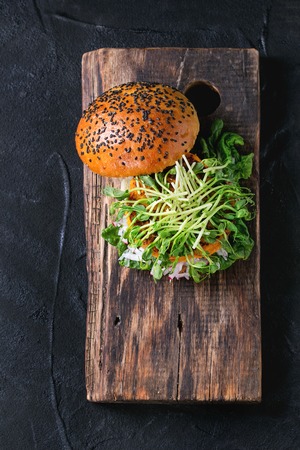 Homemade veggie sweet potato burger with fresh radish and pea sprouts served on wooden chopping board over black textured background. Top viewの写真素材