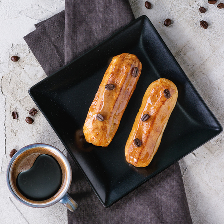 Black square plate with Coffe eclairs with cup of fresh black coffee and coffee beans on gray textile napkin over plastered surface.  Top view. Square imageの写真素材