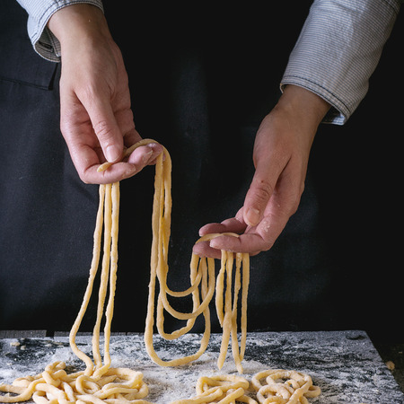Female hands making pasta pici over wooden kitchen table, powdering by flour. Dark rustic style. Square imageの写真素材