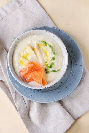 Bowl of white asparagus cream soup with pea sprouts, salted salmon and toast on gray textile napkin over white wooden background. Flat layの写真素材
