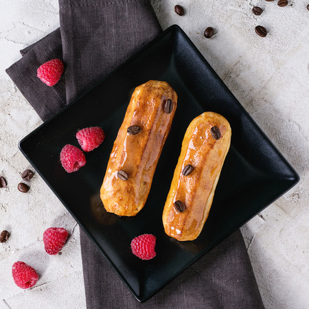 Two Coffe eclair with raspberries and coffee beans on black square plate with dark textile napkin...