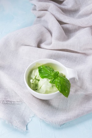 Homemade basil ice cream with fresh basil leaves in white ceramic bowl on white textile rag over blue textured background.の写真素材