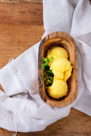 Homemade mango ice cream with fresh mint in olive wood bowl, served on white textile napkin over wooden textured background. Flat layの写真素材