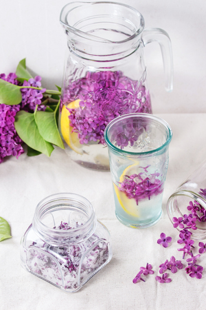 Glass jar of lilac flowers in sugar, glass and pitcher of lilac water with lemon and branch of fresh lilac on white linen tablecloth.の写真素材