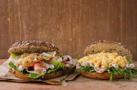 Two Whole Grain bagels with fried onion, scrambled eggs, green salad and prosciutto ham on paper over wooden textured background.の写真素材