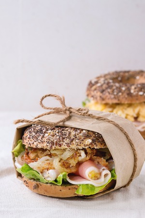 Whole Grain papered bagels with fried onion, scrambled eggs, green salad and prosciutto ham over white linen tablecloth.の写真素材