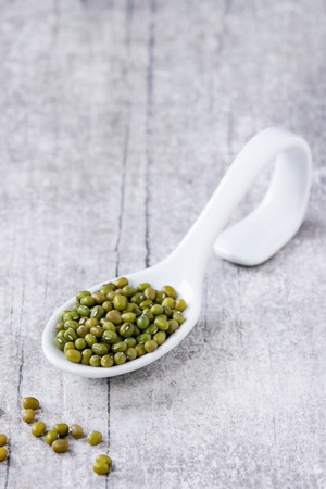 Healthy superfood. Uncooked green mungo beans in white ceramic spoon over white wooden background.の写真素材