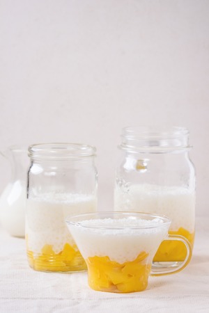 Healthy tapioca pearls pudding dessert with coconut milk and mango. Served in glass jars with jug of milk on over white linen tablecloth with white background.の写真素材