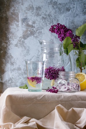 Glass jar of lilac flowers in sugar, glass and pitcher of lilac water with lemon, and branch of fresh lilac on white linen tablecloth with blue textured wall at background.の写真素材