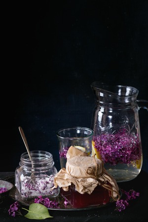 Glass jar of homemade lilac syrup, glass jar of sugared lilac flowers and glass pitcher of lilac lemonade water on black tablecloth over black. Dark rustic atmosphere.の写真素材