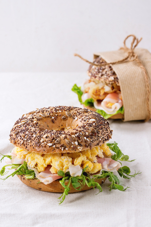 Whole Grain bagels with scrambled eggs, pea sprout and prosciutto ham on wood plate over wooden textured background.の写真素材