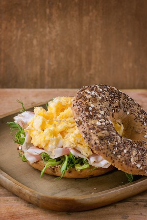 Opened Whole Grain bagels with scrambled eggs, pea sprout and prosciutto ham on wood plate over wooden textured background.の写真素材