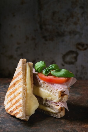 Whole grain grilled sandwich bread with melting hot cheese, ham, tomato and basil over dark rusty iron textured background. With copy spaceの写真素材