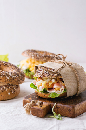 Empty and staff sprinkle seeds Whole Grain bagels with scrambled eggs, pea sprout, fried onions and prosciutto ham, making on white linen tablecloth and small wooden chopping board.の写真素材